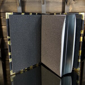May include: An open book with textured pages in shades of grey and brown. The book has a black spine and gold-coloured accents on the edges. The book is displayed on a reflective surface.