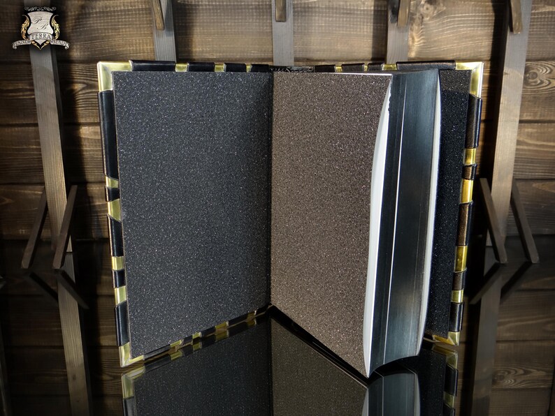 May include: An open book with textured pages in shades of grey and brown. The book has a black spine and gold-coloured accents on the edges. The book is displayed on a reflective surface.