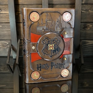 Può includere: Un libro rilegato in pelle marrone con il titolo "Harry Potter e la Pietra Filosofale". La copertina presenta immagini in rilievo del castello di Hogwarts e lo stemma di Hogwarts, con dettagli rossi. Sono visibili anche monete decorative.