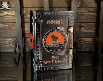 May include: A black leather-bound book titled The Odyssey by Homer, featuring a central circular orange inlay depicting a black ship on waves. The cover is embossed with a Greek key border pattern and includes two horizontal black leather straps.