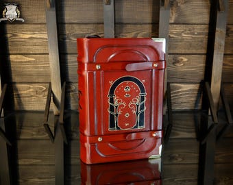 May include: A red leather book-shaped box with gold detailing and a black and gold design. The rectangular box has a handle. The background is a wooden surface.