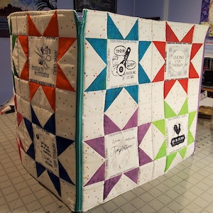 May include: A white fabric storage box with a zipper closure. The box is decorated with a patchwork design of colourful stars and squares. Each star and square has a different design and text, including "Stitches Quilt Shop", "Quilting is my therapy", and "Heavenly Stitches".