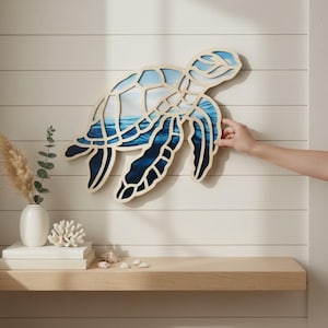 May include: A layered wooden wall art piece shaped like a sea turtle, featuring a cutout design that reveals a printed ocean scene with gradients of light blue, turquoise, and deep navy blue colors, mounted on a white horizontal paneled wall.