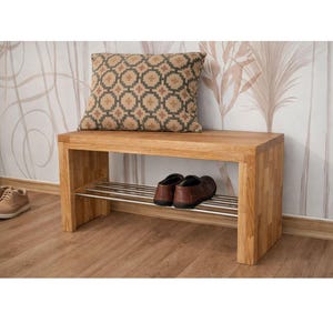 May include: A rectangular light brown wooden bench with a flat top and a lower metal shelf. A square throw cushion with a repeating geometric pattern in brown and beige sits on the bench. A pair of brown shoes rests on the metal shelf below.