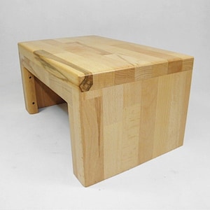 Pode incluir: Um banquinho de madeira de cor clara com acabamento natural. O banquinho é feito de várias peças de madeira que se juntam para criar um móvel resistente e durável.