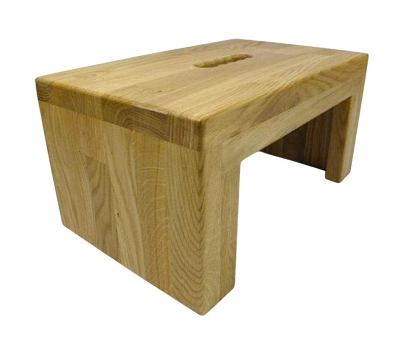 K&ouml;nnte beinhalten: Ein helles Holztrittschemel mit rechteckiger Oberseite und einem ausgeschnittenen Griff. Der Hocker hat ein schlichtes, modernes Design mit einem stabilen Sockel. Die Holzmaserung ist sichtbar und verleiht ihm ein nat&uuml;rliches Aussehen.