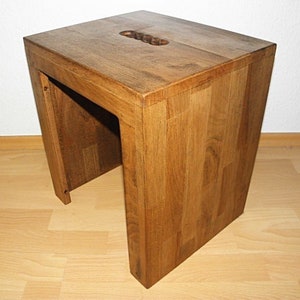 Op de afbeelding: Een houten kruk met een rechthoekig bovenblad en een uitsparing in het midden. De kruk is gemaakt van lichtbruin hout en heeft een natuurlijke afwerking.