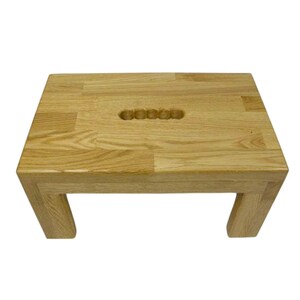 K&ouml;nnte beinhalten: Ein kleiner, rechteckiger Holzschemel mit nat&uuml;rlicher Oberfl&auml;che. Der Schemel hat einen eingelassenen Griff auf der Oberseite und vier stabile Beine. Die Holzmaserung ist im gesamten Schemel sichtbar.