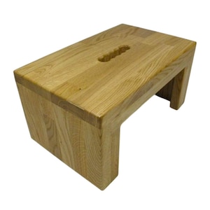 K&ouml;nnte beinhalten: Ein helles Holz-Trittschemel mit rechteckiger Oberseite und Griffaussparung. Der Hocker hat ein schlichtes, modernes Design mit abgewinkelten Beinen und ist aus Massivholz gefertigt.