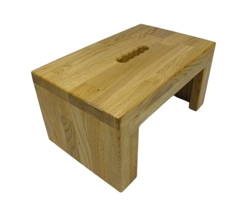 K&ouml;nnte beinhalten: Ein helles Holz-Trittschemel mit rechteckiger Oberseite und Griffaussparung. Der Hocker hat ein schlichtes, modernes Design mit abgewinkelten Beinen und ist aus Massivholz gefertigt.