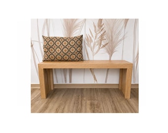 Op de afbeelding: Een rechthoekige bank van lichtgekleurd hout met een gladde afwerking en strakke lijnen. Aan de linkerkant van de bank ligt een rechthoekig sierkussen met een donkerbruin en beige geometrisch patroon met herhalende cirkelvormige motieven.