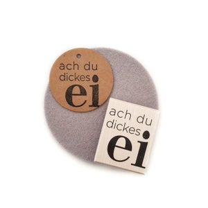 Könnte beinhalten: Zwei graue Filzuntersetzer mit dem Text "ach du dickes ei" darauf gedruckt. Ein Untersetzer hat den Text auf einem braunen Papieretikett, der andere Untersetzer hat den Text direkt auf den Filz gedruckt.