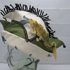 Könnte beinhalten: Eine Narrenkappe aus cremefarbenem Stoff mit blauem Dinosaurier-Druck und olivgrünen Einsätzen. Die Kappe ist mit schwarzer Fransenborte, goldfarbenem Metallbesatz, einer kleinen goldenen Glocke an der Spitze und einer Alligator-Figur verziert.