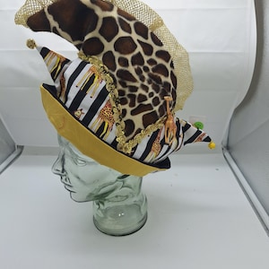 Könnte beinhalten: Eine Narrenkappe mit einem braun-cremefarbenen Giraffenmuster oben, einem schwarz-weiß gestreiften Band mit Giraffenillustrationen, einem goldenen Netz-Kamm, goldener Paillettenborte und kleinen goldenen Glöckchen an den Spitzen. Auf einem transparenten Kopf.