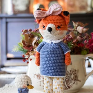 May include: A handmade crochet fox doll with an orange and white face, wearing a blue shirt, patterned pants, and brown boots. A pink bow adorns its head. A small crochet duck with a blue scarf stands nearby. The dolls are on a white lace surface.