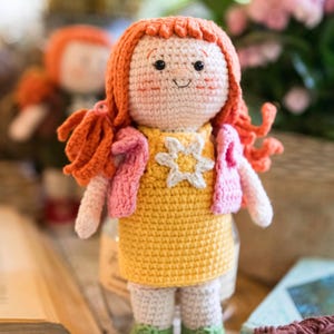 Op de afbeelding: Een handgemaakte gehaakte pop met fel oranje garen haar, gekleed in een gele jurk met een witte sterapplicatie, een roze gehaakt vestje en groene schoenen met witte veters. De pop heeft een lichtbeige huidskleur, zwarte kraaloogjes en een geborduurde glimlach.