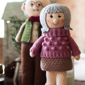 May include: Two handmade crochet dolls, a couple, stand side-by-side. The woman doll wears a pink sweater and brown skirt. The man doll wears a green vest and brown pants. Both dolls have smiling faces and are on a woven mat.
