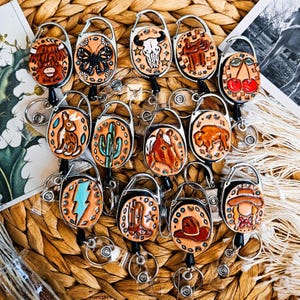 May include: A collection of round, hand-tooled leather badge reels featuring various embossed designs including a highland cow, butterfly, bull skull, cherries, rabbit, cactus, horse, buffalo, lightning bolt, cowboy boots, hat, and a person in a hat.