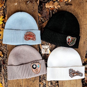 May include: Four ribbed knit beanies in light blue, black, taupe, and white. Each beanie has a leather patch with designs including a lobster, cactus, 8-ball, and a rabbit. The hats are displayed on a wooden surface with autumn leaves.