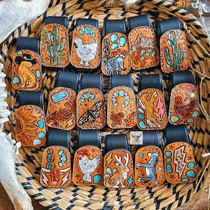 May include: A collection of seventeen rectangular leather belt clips featuring hand-tooled designs including cacti, animals, butterflies, and lightning bolts. Each piece has a black leather strap attachment and tan leather with painted accents.