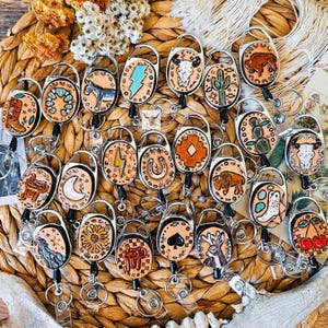 May include: A collection of oval-shaped leather badge reels featuring various hand-tooled designs including cacti, skulls, lightning bolts, animals, and floral patterns. Each badge reel has a metal clip and a retractable cord with a key ring.