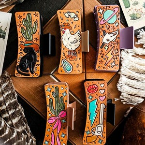 May include: Five rectangular hair claw clips featuring tan surfaces with various colorful illustrations including a black cat with a cactus, a chicken in boots, a planet, a cactus with a pink bow, and a revolver with a lightning bolt and lips.