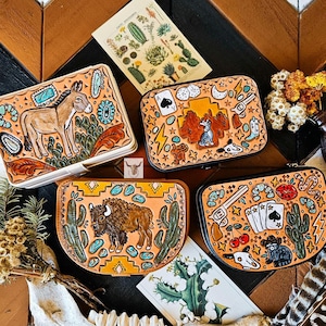 May include: Four rectangular leather cases featuring hand-tooled designs. One shows a donkey, another a buffalo, and two feature western motifs including playing cards, cacti, revolvers, boots, and skulls, all accented with turquoise-colored details.