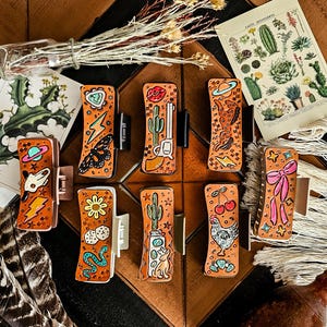 May include: Eight rectangular hair clips with tan leather-like surfaces, each featuring unique embossed and painted designs including cacti, a revolver, a butterfly, a skull, a chicken, cherries, a bow, and various celestial symbols like stars and planets.