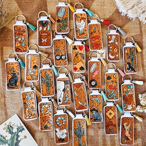 May include: A collection of rectangular tan leather keychains featuring embossed and hand-painted designs including cacti, animals, boots, and playing card symbols. Each keychain is attached to a metal ring with a small colourful tassel.
