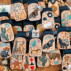 Peut inclure: Une collection de porte-clés en cuir rectangulaires présentant des motifs gravés à la main, notamment des papillons, des cactus, des animaux et des symboles de cartes à jouer. Chaque pièce a une lanière en cuir noir et des accents colorés.