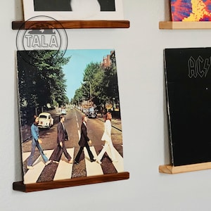 May include: Wooden record album display shelves. The shelves are made of dark wood and are mounted on a white wall. One shelf displays the album cover of The Beatles' "Abbey Road". Other album covers are also displayed.