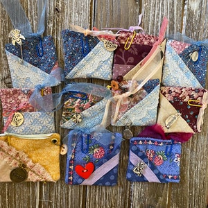 May include: A collection of small, handmade fabric pouches in various colors and patterns. Each pouch is adorned with lace, ribbons, charms, and paper clips. The pouches feature floral and geometric designs, with a rustic, vintage aesthetic.
