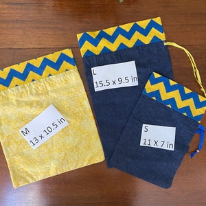 May include: Three fabric drawstring bags in different sizes. The bags are made of blue denim with yellow chevron trim. The bags are labeled with their size and dimensions in inches: Small (11 x 7 in), Medium (13 x 10.5 in), and Large (15.5 x 9.5 in).