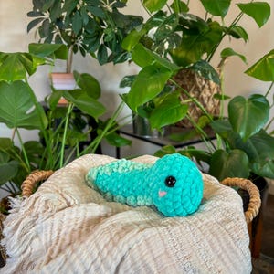 May include: A teal blue crocheted plush toy shaped like a caterpillar with a cute face. The toy is lying on a white blanket in a wicker basket.