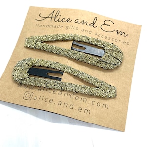 以下が含まれることがあります： 茶色のカードに、ゴールドのグリッターヘアクリップが2つ。カードには「Alice and Em Handmade gifts and Accessories www.aliceandem.com @alice.and.em.」の文字が書かれている。