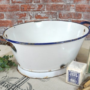 Könnte beinhalten: Weiße Emaille-Schüssel mit blauem Rand und zwei Griffen. Die Schüssel hat eine leichte Rostpatina und steht auf einer weißen Oberfläche. Im Hintergrund befinden sich Holz-Wäscheklammern und eine Schachtel Waschmittel.