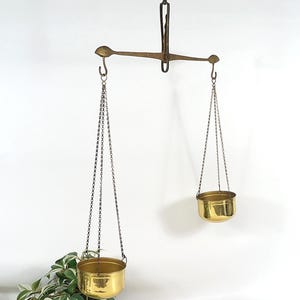 Op de afbeelding: Een hangende koperen balansweegschaal met een centrale horizontale balk die aan een haak is bevestigd. Twee gepolijste koperen schalen zijn met dunne metalen kettingen aan de uiteinden van de balk bevestigd. De weegschaal is asymmetrisch.