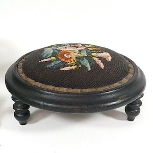 Peut inclure: Un repose-pieds rond avec un cadre en bois noir et quatre pieds courts tournés. Le dessus est recouvert d'un tissu marron foncé avec un motif floral central en point de croix dans des tons orange, rose, blanc et vert, bordé d'une garniture.
