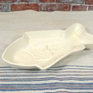 Op de afbeelding: Off-white keramische visvormige serveerschaal. De schaal heeft een textuur met schubbenpatroon en een rechthoekig compartiment. De schaal ligt op een beige en blauw gestreepte stof.