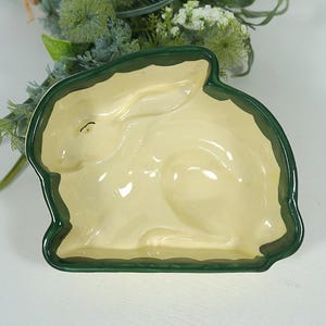 Könnte beinhalten: Eine keramische, hasenförmige Schale mit cremefarbenem Innenraum und dunkelgrünem Rand. Die Schale hat ein detailliertes Relief eines Kaninchens und dient wahrscheinlich zum Servieren oder Dekorieren. Der Hintergrund enthält Grün.