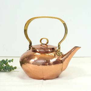 Op de afbeelding: Een koperen theepot met een gehamerde afwerking. Het heeft een messing handvat en tuit, en een geweven rotan grip op het handvat. Het deksel heeft een kleine messing knop. De ketel heeft een warme koperkleur.
