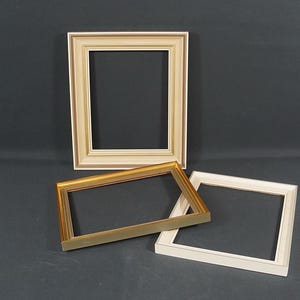 Könnte beinhalten: Drei rechteckige Bilderrahmen sind abgebildet. Ein Rahmen ist vertikal ausgerichtet und hat ein cremefarbenes Finish, während zwei Rahmen horizontal liegen, einer mit einem metallisch goldenen Finish und der andere in Creme mit goldenen Akzenten.