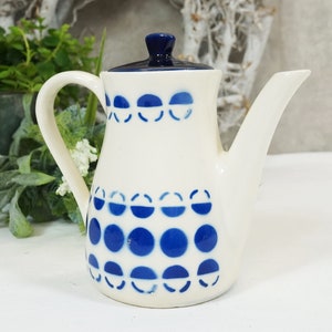 Op de afbeelding: Een witte theepot van keramiek met een blauw deksel en een blauw-wit geometrisch patroon. De theepot heeft een rond lichaam en een tuit aan de ene kant.