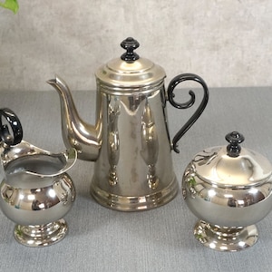 Könnte beinhalten: Ein silbernes Kaffee-Set mit Kanne, Milchkännchen und Zuckerdose mit schwarzen Griffen und Knöpfen. Das Set ist aus poliertem Metall und hat einen Vintage-Look.