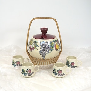 Può includere: Una ciotola per punch in ceramica bianca con un coperchio rosso e un manico in vimini. La ciotola è decorata con un motivo di frutta colorata. Sono incluse quattro tazze in ceramica abbinate con motivi di frutta.