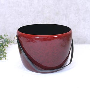 Könnte beinhalten: Ein weinroter Eimer mit schwarzem Innenraum und schwarzem Griff. Der Eimer hat eine glänzende Oberfläche und eine gesprenkelte Textur. Der Griff ist mit kleinen Metallschlaufen befestigt. Der Eimer steht auf einer weißen Oberfläche.