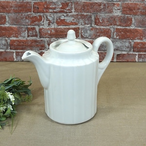 Op de afbeelding: Een witte keramische theepot met een deksel en handvat. De theepot heeft een verticaal geribbeld ontwerp en een tuit om uit te schenken. De achtergrond heeft een bakstenen muur en groen.