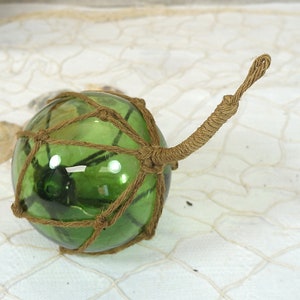 Può includere: Un galleggiante da pesca in vetro verde con un manico in corda marrone. Il galleggiante è avvolto in una rete marrone.