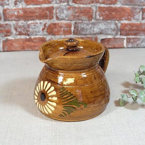 Peut inclure: Pichet en céramique marron avec couvercle et anse. Le pichet présente un motif floral avec une marguerite blanche et des feuilles vertes. Le pichet est posé sur une surface claire devant un mur de briques.