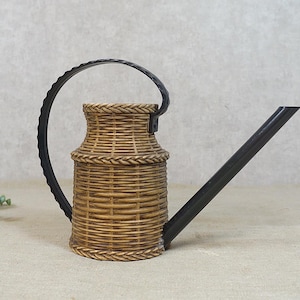 Può includere: Un annaffiatoio decorativo con un design a cestino intrecciato. Il corpo dell'annaffiatoio è color camoscio con un manico e un beccuccio neri. Il manico ha un design strutturato e scanalato. Il beccuccio è lungo e angolato.